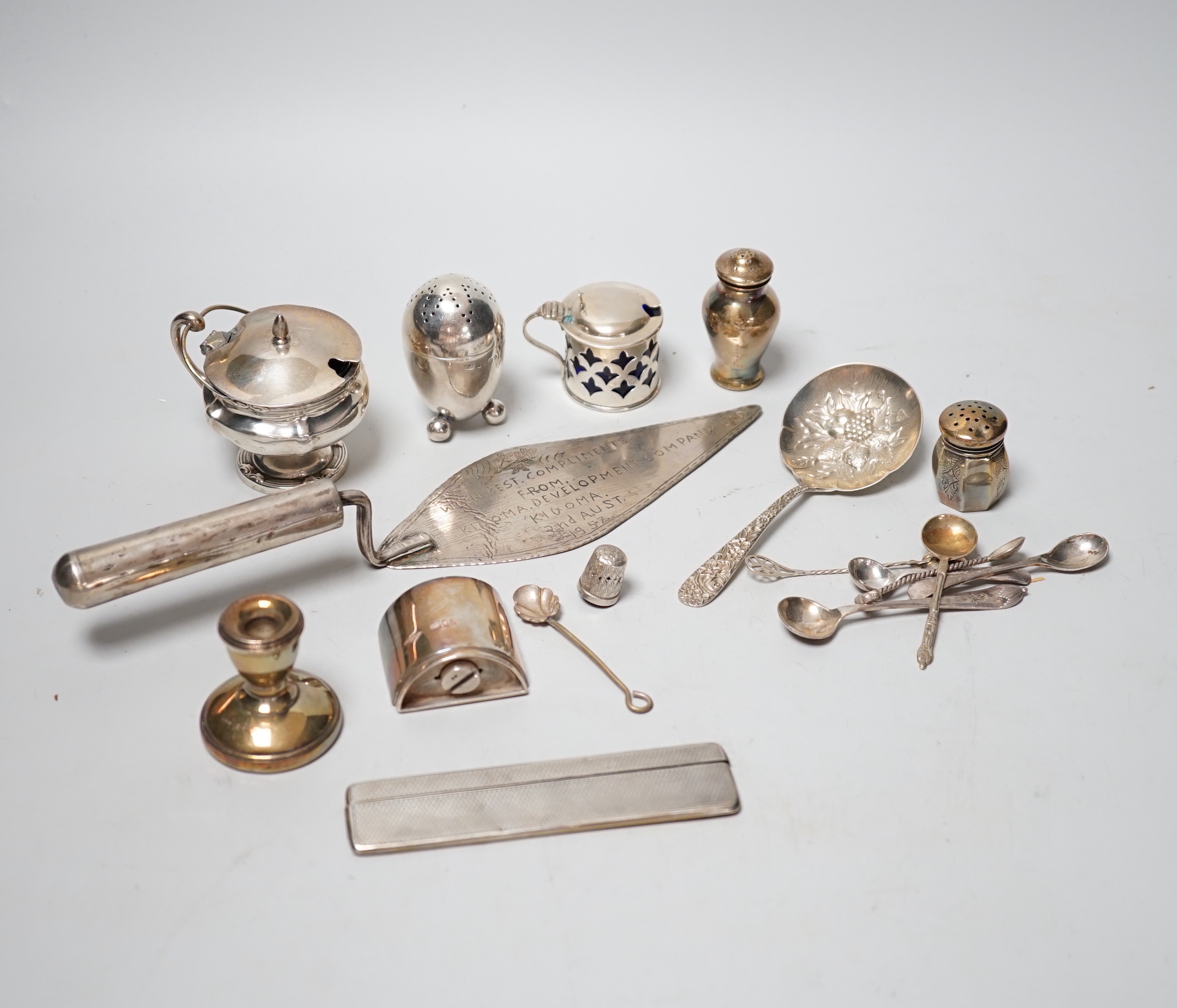 A collection of assorted small silver including condiments, spoons, egg cup, pill box, comb, Tiffany & Co pen, dwarf candlesticks, and other items including a plated evening bag and African trowel.